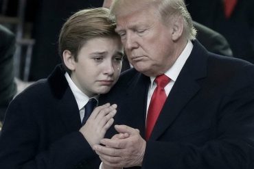Barron Trump in formal black suit standing with an adult in dark suit and red tie
