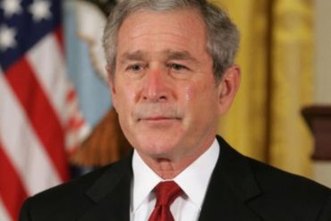 George W. Bush at World Series event wearing black suit and red tie with American flag background
