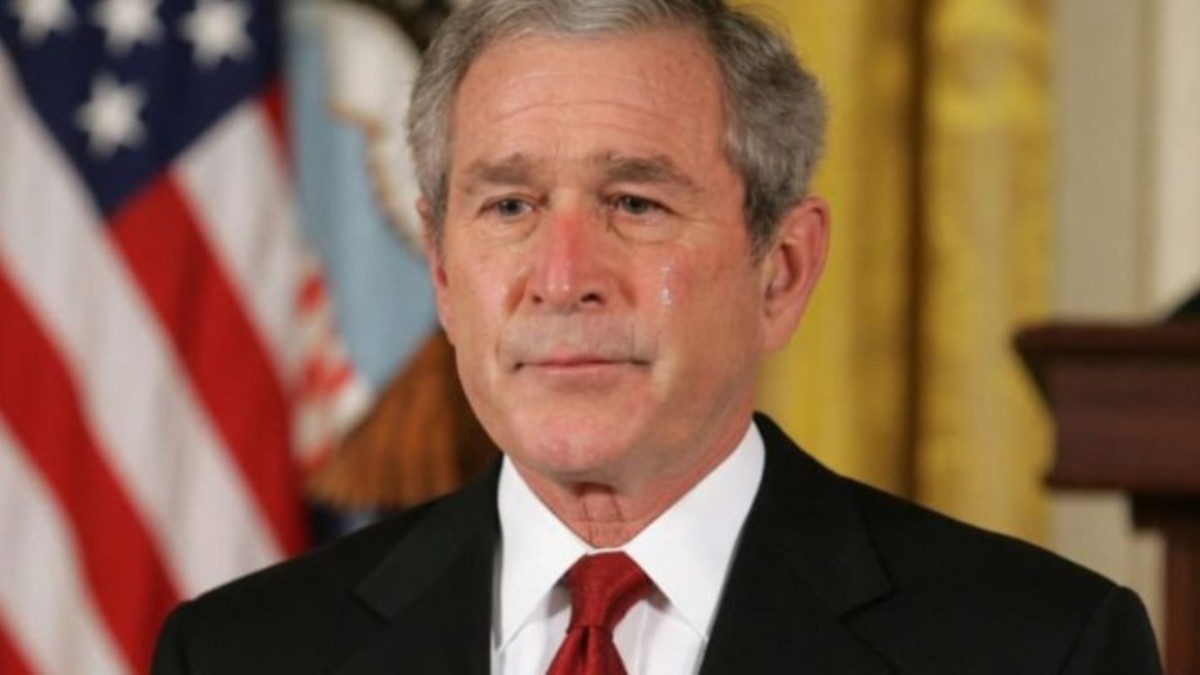 George W. Bush at World Series event wearing black suit and red tie with American flag background